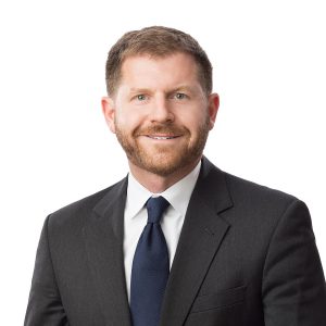 A person with a beard is wearing a dark suit with a white shirt and a blue tie against a white background.