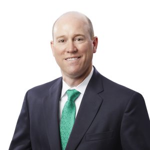 A smiling person wearing a dark suit with a green tie poses against a plain background.