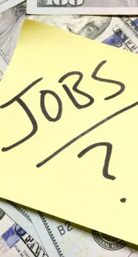 A sticky note with the word "JOBS" written on it is placed on top of a pile of U.S. hundred-dollar bills.