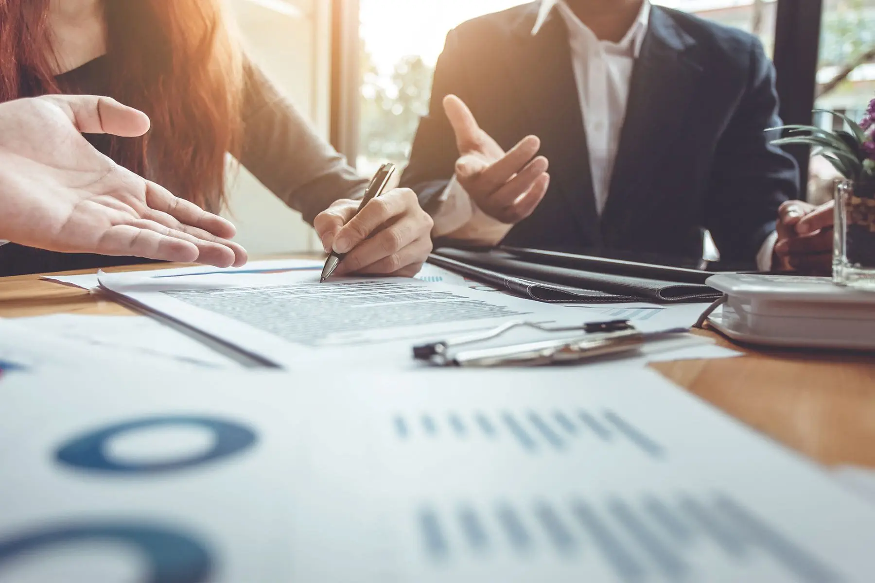 Individuals are engaged in a business meeting, with one person pointing at a document on a table scattered with various papers and reports.