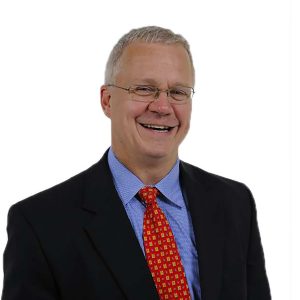 A smiling individual wearing glasses, a blue suit, and a red tie with yellow patterns against a plain white background.