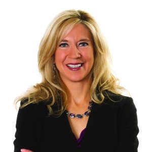 A smiling woman with blonde hair is wearing a black blazer and a purple blouse, accessorized with a multi-colored necklace.