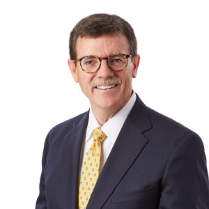 A man with glasses wearing a blue suit and a yellow tie is smiling against a plain background.