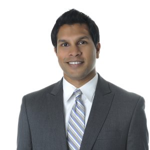A person is wearing a dark suit, light blue shirt, and a striped tie against a white background.