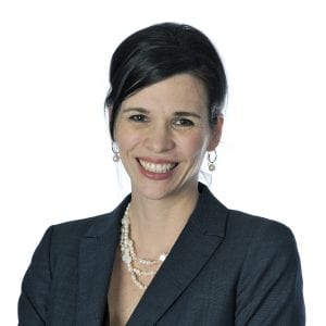 A smiling woman with a pearl necklace and earrings is wearing a dark blazer over a light-colored top with her dark hair styled in a short bob cut against a white background.